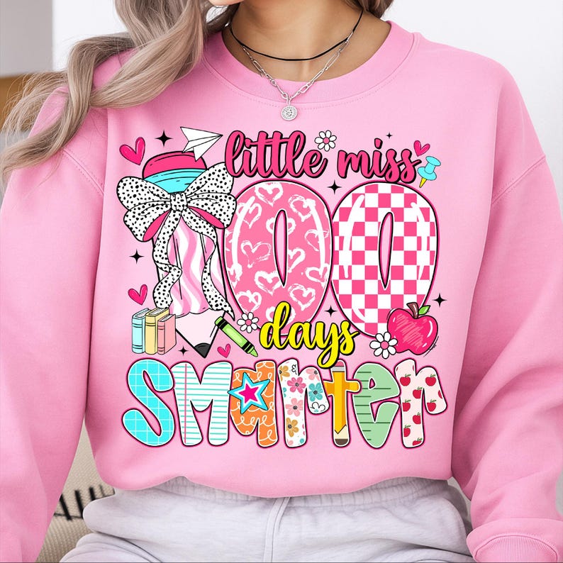 May include: Pink sweatshirt with a colorful graphic celebrating 100 days of school. The design includes the words "Little Miss 100 Days Smarter" with hearts, flowers, and an apple. A large bow and books are also featured.