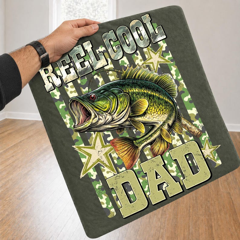 May include: A dark green blanket with the words "REEL COOL DAD" in a distressed font. The blanket features a detailed illustration of a largemouth bass with an open mouth, set against a camouflage and star design.
