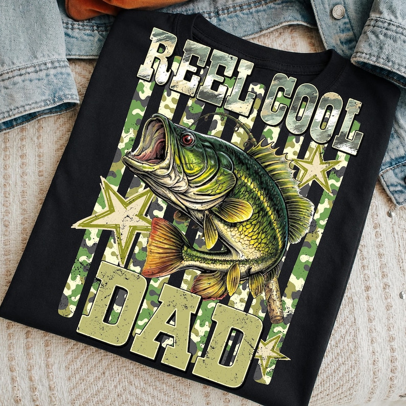 May include: Black t-shirt with a graphic of a green and yellow fish with an open mouth. The text "REEL COOL DAD" is printed above and below the fish in a camouflage font. The design includes green and white stars and stripes.