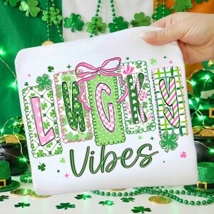Op de afbeelding: Wit T-shirt met de woorden "LUCKY VIBES" in groen en roze, versierd met klavers en een roze strik. De letters zijn gevuld met verschillende patronen. Het shirt wordt door een hand omhoog gehouden.