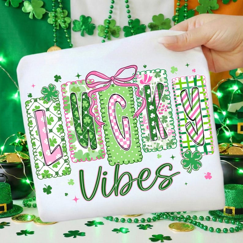 Op de afbeelding: Wit T-shirt met de woorden "LUCKY VIBES" in groen en roze, versierd met klavers en een roze strik. De letters zijn gevuld met verschillende patronen. Het shirt wordt door een hand omhoog gehouden.