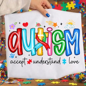 May include: White t-shirt with the word "AUTISM" in colorful, outlined letters. The shirt includes the phrases "accept," "understand," and "love" below the word. Decorative elements include hearts, flowers, stars, and puzzle pieces.