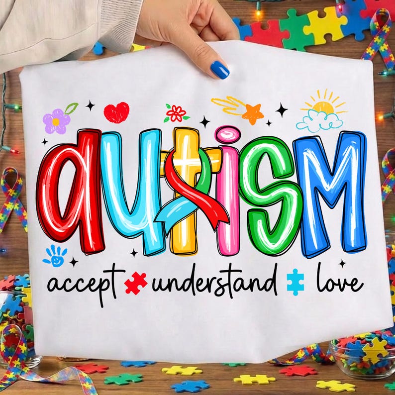 May include: White t-shirt with the word "AUTISM" in colorful, outlined letters. The shirt includes the phrases "accept," "understand," and "love" below the word. Decorative elements include hearts, flowers, stars, and puzzle pieces.