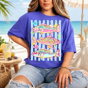 May include: A purple t-shirt featuring three colorful fish designs with floral accents and vertical blue stripes. The fish are in shades of pink, yellow, and blue. The shirt is a casual, comfortable style.