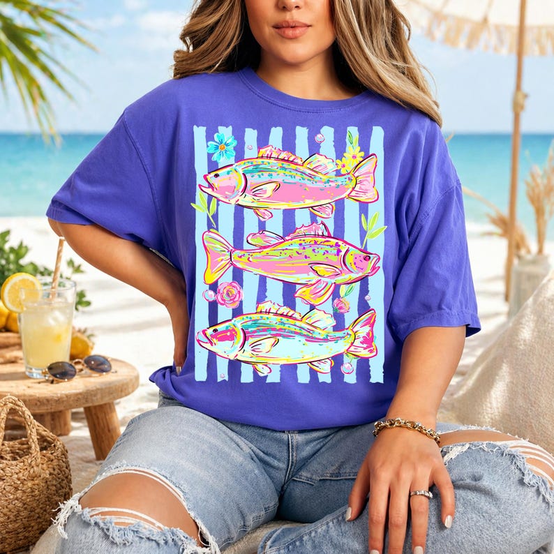 May include: A purple t-shirt featuring three colorful fish designs with floral accents and vertical blue stripes. The fish are in shades of pink, yellow, and blue. The shirt is a casual, comfortable style.
