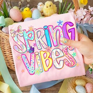 May include: A light pink t-shirt with the words "SPRING VIBES" in colorful, floral-themed lettering. The shirt is held in front of a wicker basket filled with Easter eggs and decorative chicks, with a wooden background.