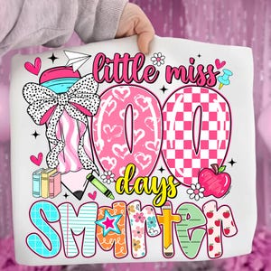 May include: A white tote bag featuring a colorful design celebrating 100 days of school. The design includes the words "little miss 100 days smarter" with hearts, flowers, and an apple.