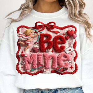 Peut inclure: Sweat-shirt blanc avec un motif de Saint-Valentin. Le motif pr&eacute;sente les mots "Be Mine" en rouge et rose, ainsi qu'un cupidon, des c&oelig;urs et un arc et une fl&egrave;che. Le motif est encadr&eacute; d'une bordure rouge.