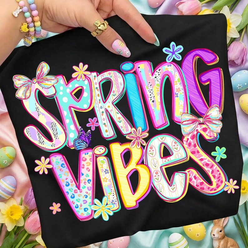 May include: Black t-shirt with the words "SPRING VIBES" in colorful, floral-themed lettering. The design includes butterflies, bows, and flowers in pastel colors.