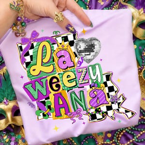 May include: A light purple t-shirt with the text "La Weezu Ana" in a colorful, layered design. The design includes a disco ball heart, a crown, and a fleur-de-lis. The shirt is surrounded by Mardi Gras beads and masks.