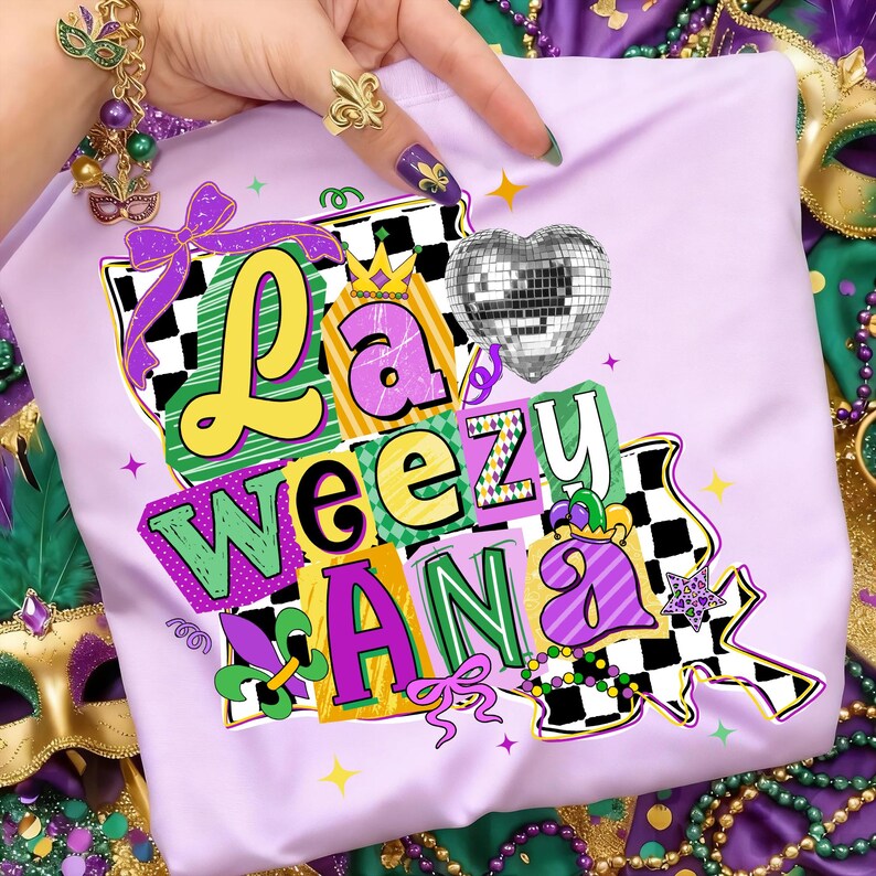 May include: A light purple t-shirt with the text "La Weezu Ana" in a colorful, layered design. The design includes a disco ball heart, a crown, and a fleur-de-lis. The shirt is surrounded by Mardi Gras beads and masks.
