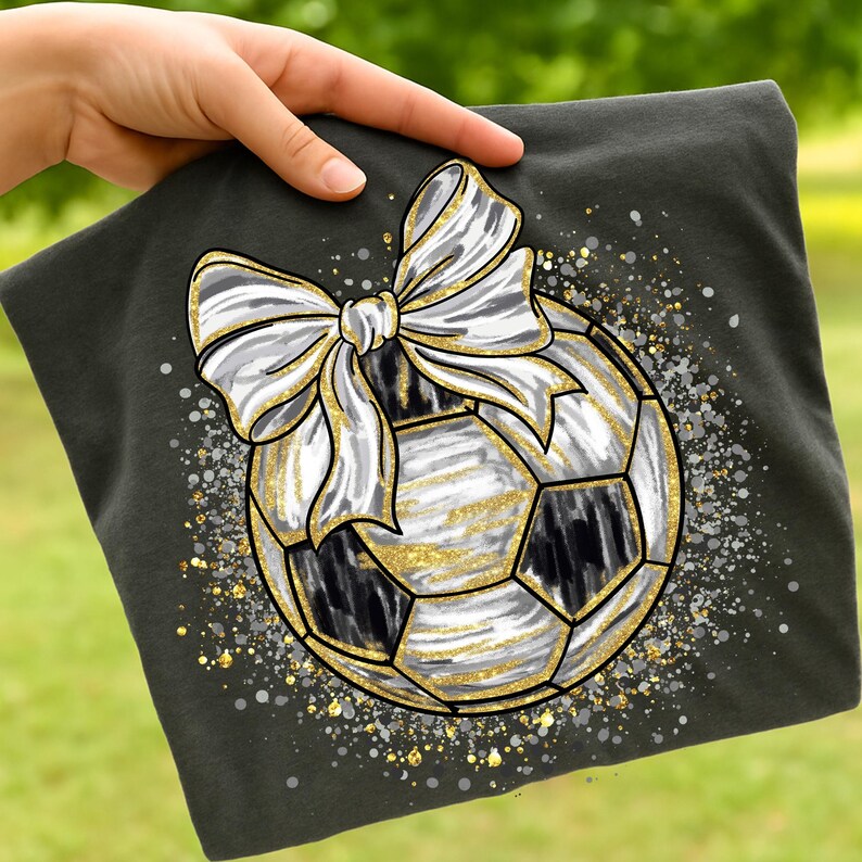 K&ouml;nnte beinhalten: Dunkelgraues T-Shirt mit einem Fu&szlig;ball-Design, einer wei&szlig;en Schleife und goldenen Glitzerakzenten. Der Fu&szlig;ball hat schwarz-wei&szlig;e Paneele mit goldenen Glitzerdetails. Das Design befindet sich vor einem Hintergrund aus kleinen, verstreuten Glitzerpartikeln.