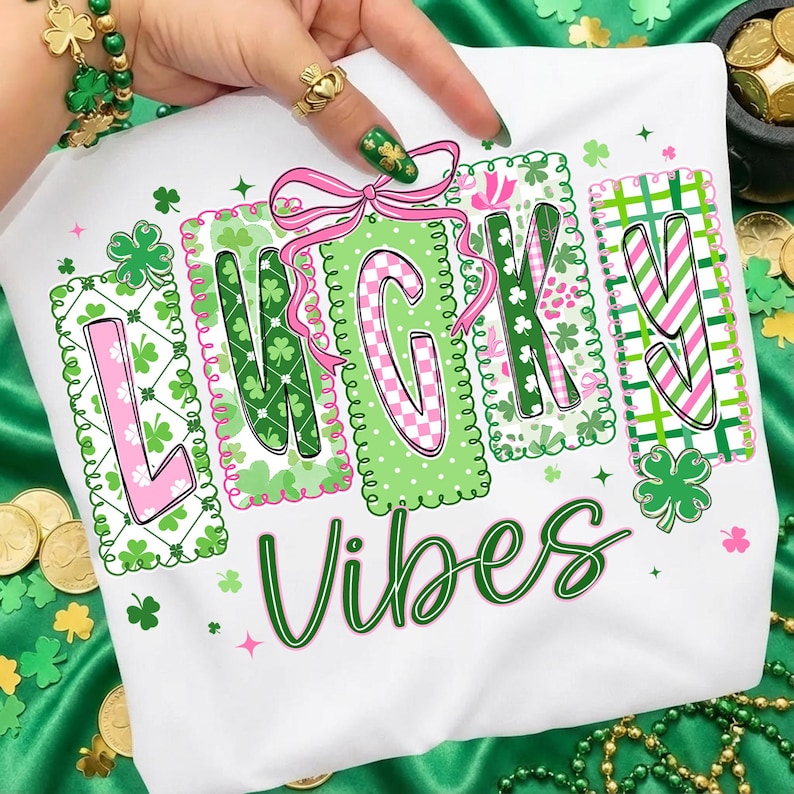 Op de afbeelding: Wit T-shirt met de woorden "LUCKY VIBES" in groen schrift. Het woord "LUCKY" staat in roze en groene blokken met klaver- en bloempatronen. Boven de letter "C" staat een roze strik. Het shirt is versierd met klavers.
