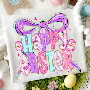 May include: White sweatshirt with a colourful "Happy Easter" graphic. The design features a large purple bow, pastel-coloured text, and Easter eggs. The text is outlined in gold glitter.
