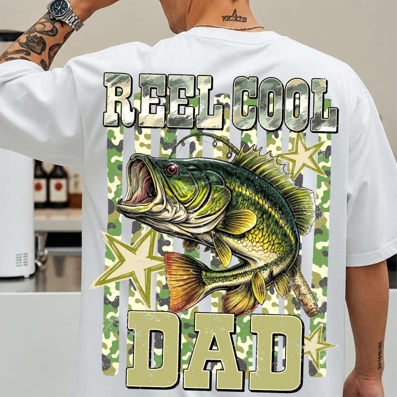 May include: White t-shirt featuring a large graphic of a green and yellow fish with an open mouth, fishing rod, and the text "REEL COOL DAD" in a camouflage design. The shirt has a classic crew neck.
