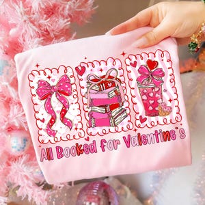 May include: A light pink sweatshirt with a Valentine's Day theme. The design features three framed illustrations: a pink bow, a stack of books, and a pink drink with hearts. The text "All Booked for Valentine's" is printed below.