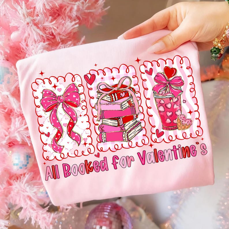 May include: A light pink sweatshirt with a Valentine's Day theme. The design features three framed illustrations: a pink bow, a stack of books, and a pink drink with hearts. The text "All Booked for Valentine's" is printed below.