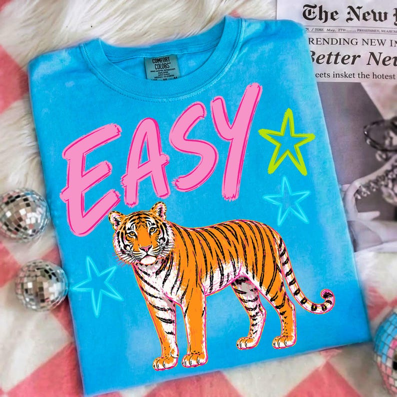 May include: A turquoise Comfort Colors t-shirt with the word "EASY" in pink block letters and a tiger graphic. Neon green and blue stars accent the design. The shirt is a casual, everyday wear item.