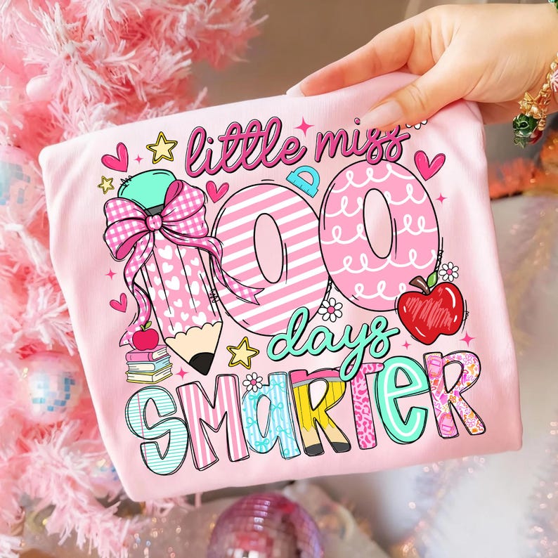 Puede incluir: Una camiseta rosa claro con la frase "little miss 100 days smarter" en una fuente colorida y juguetona. El dise&ntilde;o incluye un l&aacute;piz, corazones, estrellas y una manzana. La camiseta est&aacute; sujeta por una mano.