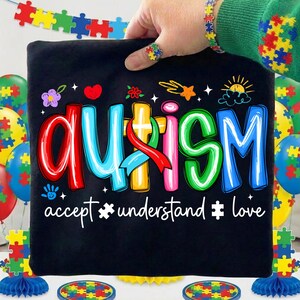 May include: Black t-shirt with the word "AUTISM" in colorful letters and the phrases "accept, understand, love." The image includes puzzle piece decorations and a matching bracelet and ring.