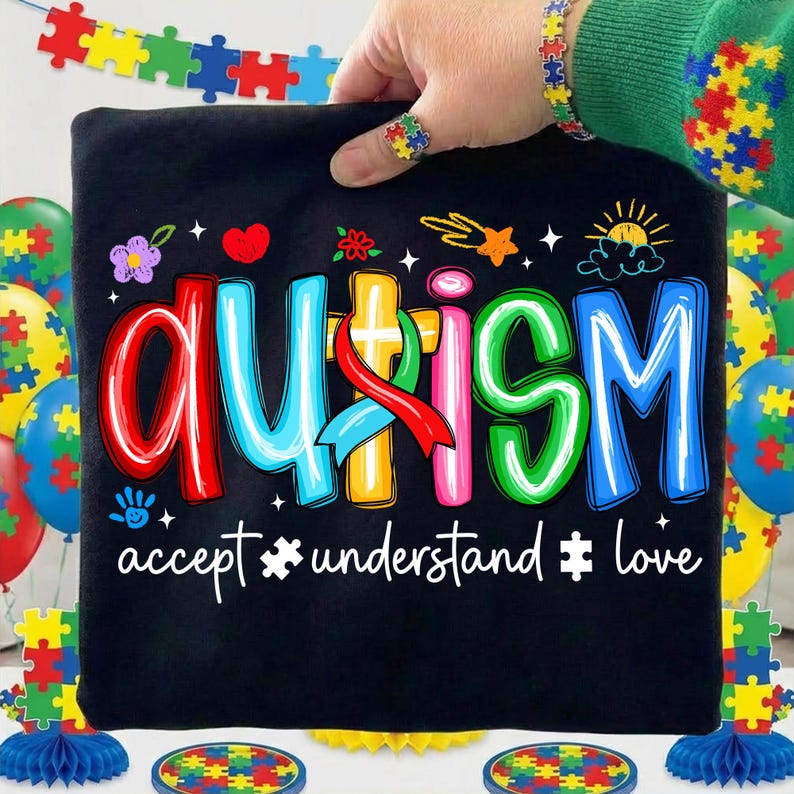 May include: Black t-shirt with the word "AUTISM" in colorful letters and the phrases "accept, understand, love." The image includes puzzle piece decorations and a matching bracelet and ring.