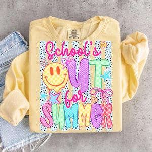 Puede incluir: Sudadera amarilla clara con el texto "School's Out for Summer" en letras coloridas y divertidas. El dise&ntilde;o incluye una carita sonriente, estrellas y flores. La sudadera es de la marca Comfort Colors.