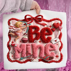 Peut inclure: Une cr&eacute;ation sur le th&egrave;me de la Saint-Valentin avec les mots "Be Mine" en rouge et rose, un cupidon, des c&oelig;urs et un n&oelig;ud. Le design est sur un fond blanc avec une bordure rouge et blanche.