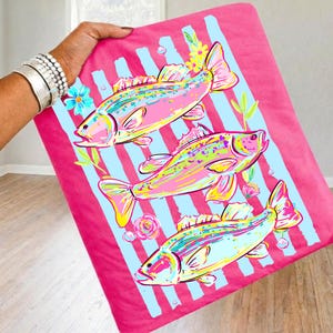 May include: A pink blanket featuring three colorful fish illustrations with blue stripes, flowers, and bubbles. The fish are in shades of pink, yellow, and blue. The blanket is held up by a person.