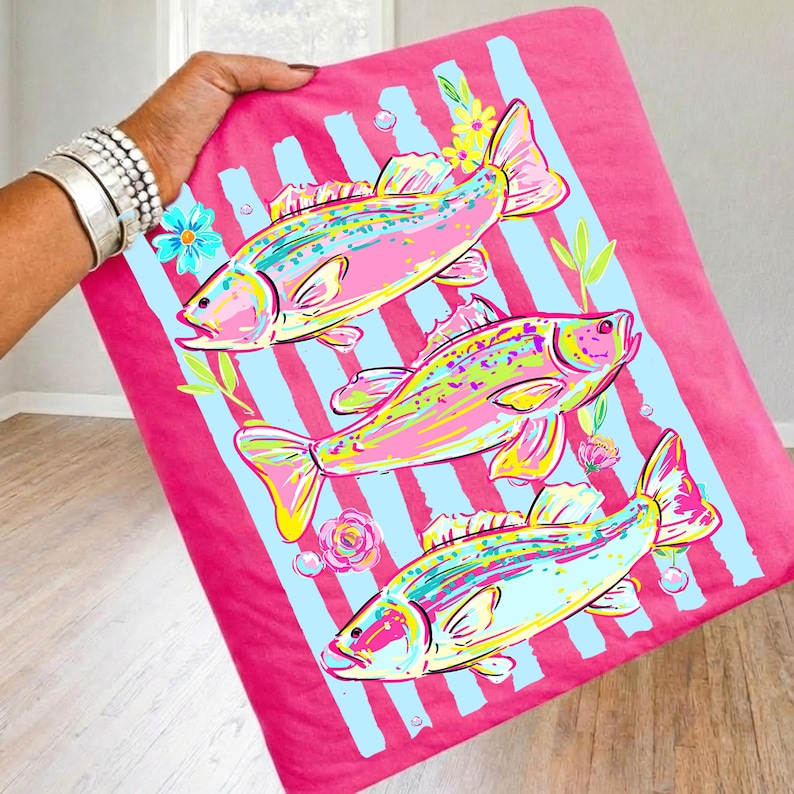 May include: A pink blanket featuring three colorful fish illustrations with blue stripes, flowers, and bubbles. The fish are in shades of pink, yellow, and blue. The blanket is held up by a person.