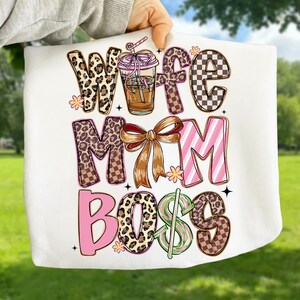 May include: White tote bag with the words "Wife Mom Boss" in a colorful design. The design includes leopard print, checkered patterns, and a coffee drink illustration. The bag is held by a person.