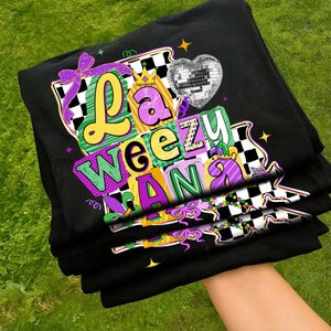 May include: Black t-shirts stacked, featuring a colorful graphic design. The design includes the words "La Weeziana" in various fonts and colors, a disco ball heart, and Mardi Gras-themed elements like a crown and beads.