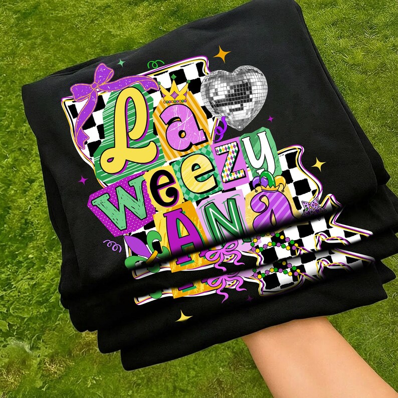 May include: Black t-shirts stacked, featuring a colorful graphic design. The design includes the words "La Weeziana" in various fonts and colors, a disco ball heart, and Mardi Gras-themed elements like a crown and beads.