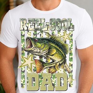 May include: White t-shirt with a graphic design featuring a green and yellow fish with an open mouth, the words "REEL COOL DAD" in a camouflage font, and star accents. The shirt is made of a soft material.