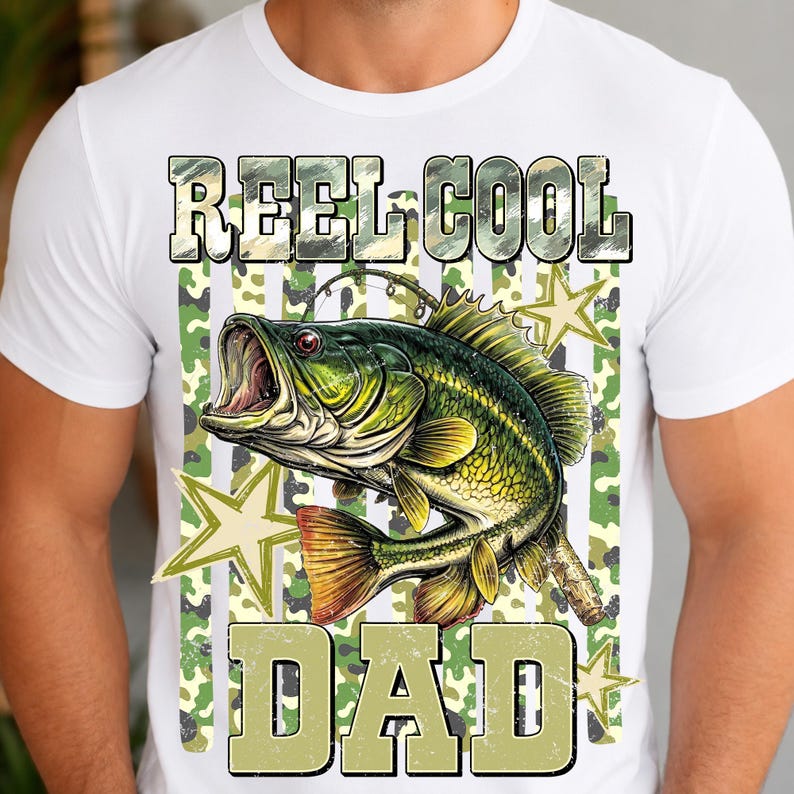 May include: White t-shirt with a graphic design featuring a green and yellow fish with an open mouth, the words "REEL COOL DAD" in a camouflage font, and star accents. The shirt is made of a soft material.