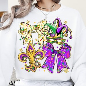 May include: White sweatshirt featuring a colorful Mardi Gras design. The design includes a fleur-de-lis, bows, a jester hat, and a mask in purple, green, and gold. The bows have decorative patterns and the sweatshirt has a crew neck.