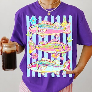 May include: Purple t-shirt featuring a colorful design of three fish with floral accents and light blue vertical stripes. The fish are in shades of pink, yellow, and green. The shirt has a round neckline and short sleeves.
