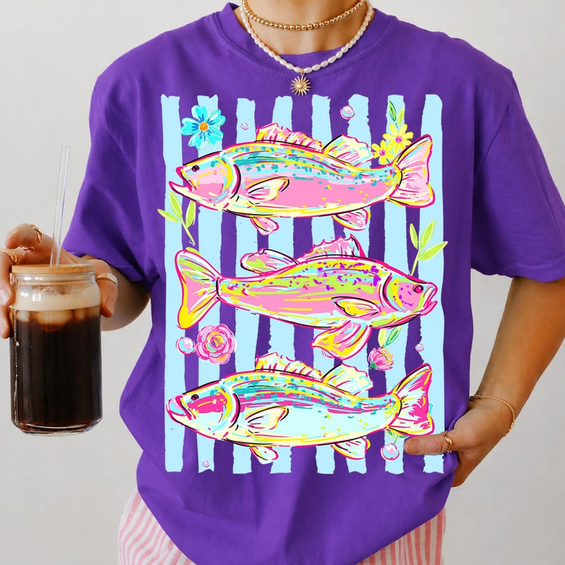 May include: Purple t-shirt featuring a colorful design of three fish with floral accents and light blue vertical stripes. The fish are in shades of pink, yellow, and green. The shirt has a round neckline and short sleeves.