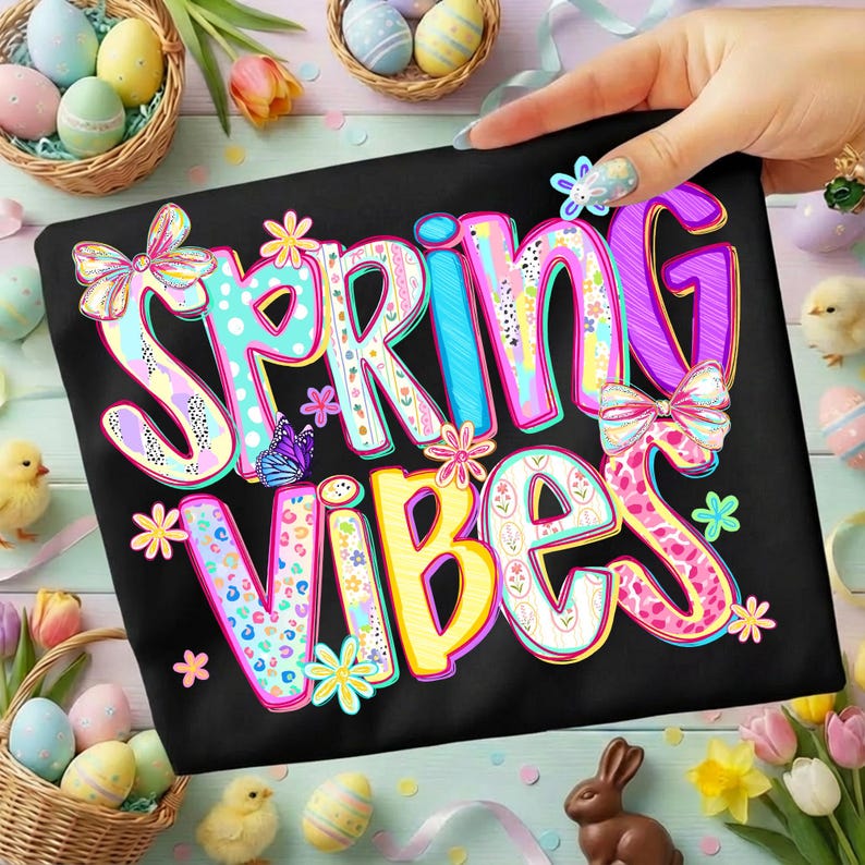 May include: Black t-shirt with the words "SPRING VIBES" in colorful, patterned letters. The design includes butterflies, flowers, and bows, with a spring theme. Easter eggs and chicks are in the background.