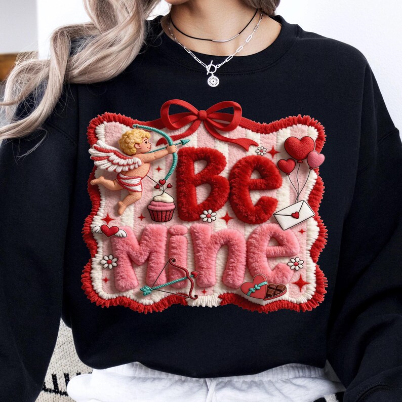 Peut inclure: Sweat-shirt noir avec un motif de Saint-Valentin. Le motif pr&eacute;sente les mots "Be Mine" en rouge et rose, un cupidon avec un arc et des fl&egrave;ches, des c&oelig;urs et un cupcake. Le motif est encadr&eacute; d'une bordure rouge et blanche.