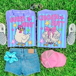 May include: A flat lay featuring a purple t-shirt with the text "GUESS WHAT" and a cartoon chicken wearing sunglasses, and another shirt with the text "CHICKEN BUTT". Also included are denim shorts, a pink trucker hat, a blue scrunchie, and checkered slip-on shoes.