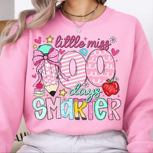 Puede incluir: Sudadera rosa con el texto "little miss 100 days smarter". El dise&ntilde;o incluye un l&aacute;piz, el n&uacute;mero 100, una manzana, estrellas y corazones. La sudadera es de cuello redondo.