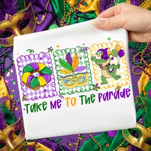 Könnte beinhalten: Weißes T-Shirt mit einem farbenfrohen Mardi Gras-Design. Das Design umfasst einen King Cake, eine Maske und ein Cartoon-Alligator, der Luftballons hält. Der Text "Take Me To The Parade" ist in Grün und Lila geschrieben.