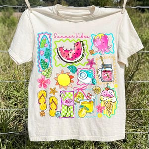 Puede incluir: Camiseta color crema con un gr&aacute;fico colorido de tem&aacute;tica veraniega. El dise&ntilde;o incluye una rodaja de sand&iacute;a, un pulpo, un cono de helado y el texto "Summer Vibes". La camiseta est&aacute; colgada en un tendedero.