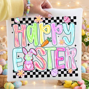 Może przedstawiać: Biała kwadratowa poduszka z napisem "Happy Easter" w kolorowych literach bąbelkowych. Projekt zawiera kr&oacute;lika, marchewkę, kwiaty i pisanki, wszystko na czarno-białej szachownicy.