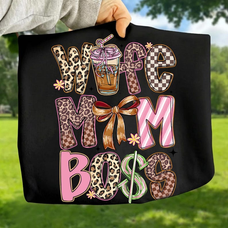 May include: Black tote bag with the words "Wife, Mom, Boss" in a mix of leopard print, checkered, and pink lettering. A coffee drink illustration is also featured. The bag is held up against a green background.