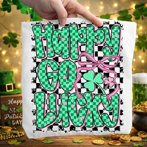 May include: White tote bag with the text "Happy Go Lucky" in green and pink, with a checkered background and shamrock accents. The bag is held up by a hand, with St. Patrick's Day decorations in the background.