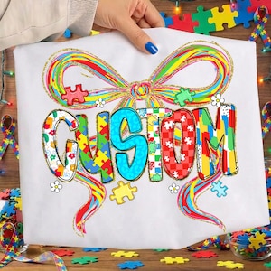 May include: White t-shirt featuring the word "CUSTOM" in colorful puzzle piece letters, with a rainbow bow design above. The design includes puzzle pieces and small white flowers. The shirt is held by a person with blue nail polish.