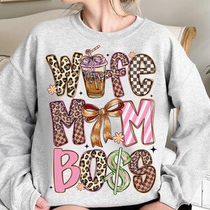 May include: A light gray sweatshirt with the words "Wife Mom Boss" in a mix of leopard print, checkered, and striped patterns. A coffee drink graphic is in the center, with a bow and floral accents.