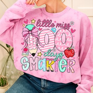 Puede incluir: Sudadera rosa te&ntilde;ida con un gr&aacute;fico colorido. El gr&aacute;fico incluye el texto "little miss 100 days smarter" con un l&aacute;piz, corazones, estrellas y una manzana. La sudadera es de cuello redondo.