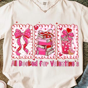 May include: Cream-colored t-shirt with a Valentine's Day graphic. The graphic features a pink bow, a stack of books, and a pink drink, all within decorative frames. The text "All Booked for Valentine's" is printed below.